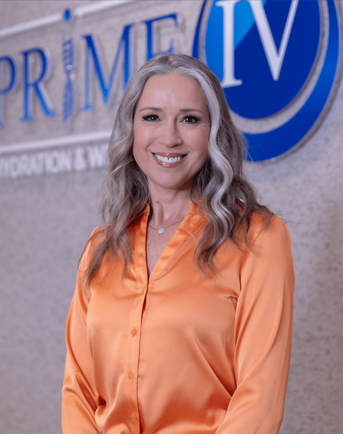 Gina Van Luven, owner of Prime IV Hydration & Wellness Mesa, smiling in front of the Prime IV logo wall inside her spa