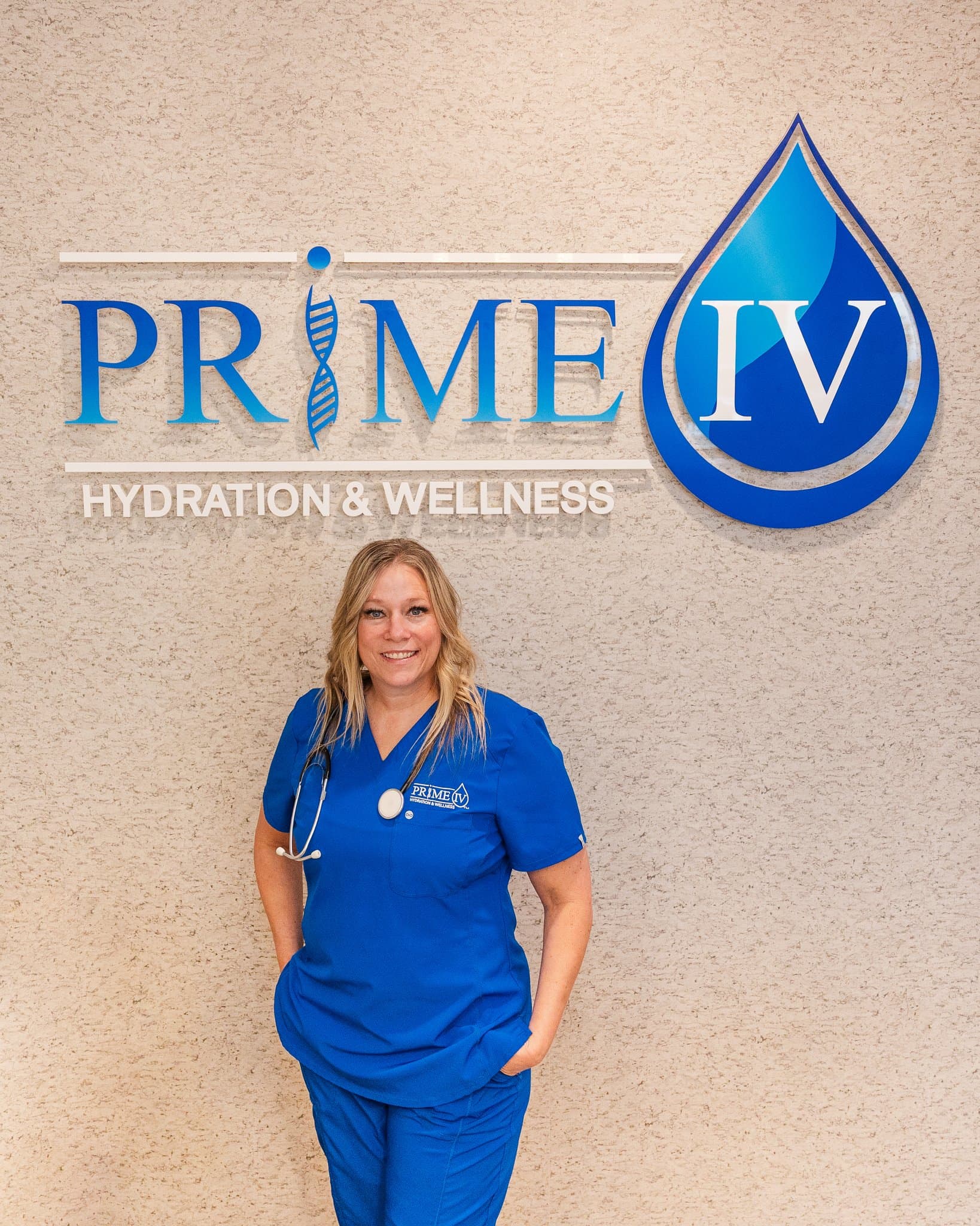 Tiffany Kuehn, Mobile & Events Director at Prime IV Mesa, in Prime IV scrubs with a stethoscope in front of the Prime IV Hydration & Wellness logo wall